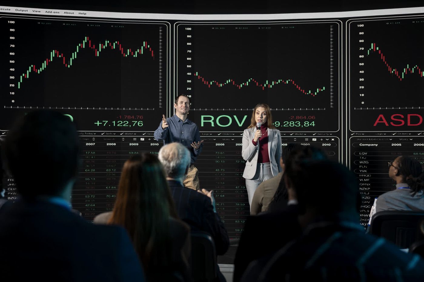 Presenters on stage in front of performance charts, speaking to a full audience
